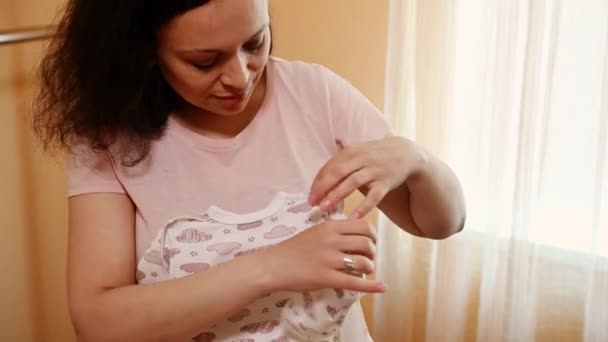 Detailní záběr okouzlující rozkošné multietnické těhotné ženy skládající oblečení pro nové dítě, stojící u žehlícího prkna, připravující tašku pro porodní nemocnici. Těhotenství. Mateřství. mateřská dovolená - Záběry, video
