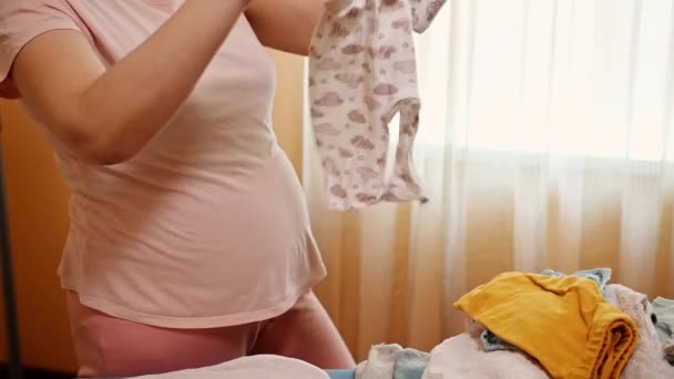 Müstakbel annenin, yeni doğan bebek için ütü masasında temiz çamaşır ve ütülü giysiler düzenlemesine yakın çekim. Hamile kadın doğum hastanesi için çamaşır hazırlıyor. Hamilelik. Annelik - Video, Çekim