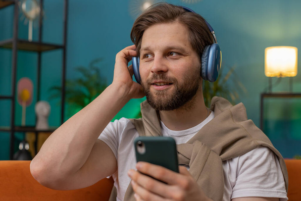Gelukkig dolblij Kaukasische man in draadloze koptelefoon ontspannen zit op de bank thuis appartement kiezen luisteren favoriete energetische disco dansen muziek. Vrijetijdsbesteding overdag - Foto, afbeelding