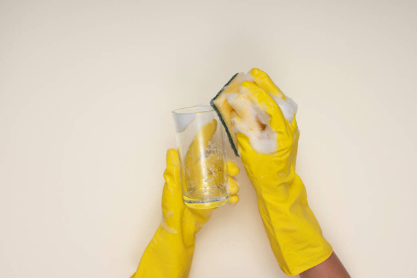 woman wearing yellow cleaning gloves on light color background cleaning glass cup with cleaning sponge - Foto, Imagem