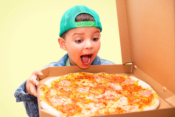 Ein siebenjähriger Junge mit grüner Mütze und hellen Schreien hält schockiert eine offene Pizzaschachtel in der Hand.. - Foto, Bild