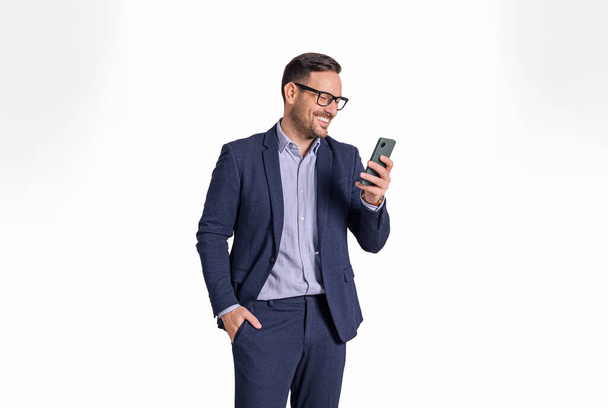 Handsome male professional manager with hand in pocket messaging online on mobile phone. Elegant businessman reading e-mails on cellphone while standing isolated over white background - Foto, Imagen