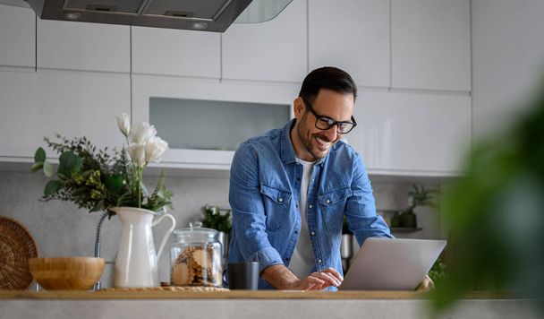 Cheerful young businessman dressed in denim shirt sending reports over laptop while standing at kitchen island. Happy male entrepreneur surfing net over computer while working from home office - Photo, Image
