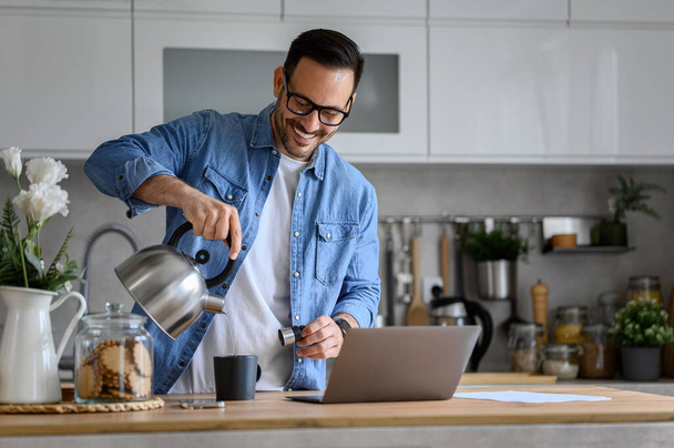 Hymyilevä nuori liikemies kaataa kuumaa kahvia kuppiin vedenkeittimestä työskennellessään kannettavan keittiön pöydällä. Iloinen mies freelancer juominen työskennellessään etänä kotitoimistosta - Valokuva, kuva