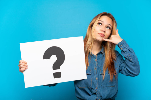 Teenager girl over isolated blue background holding a placard with question mark symbol and doing phone gesture - Фото, зображення