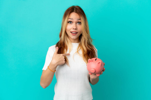 Teenager blonde girl holding a piggybank over isolated blue background with surprise facial expression - Photo, Image
