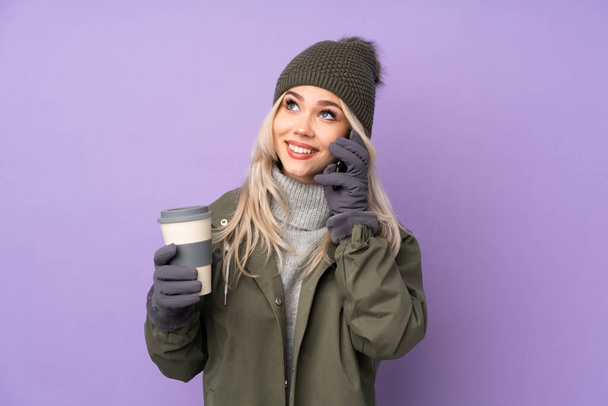 Teenager blonde girl with winter hat over isolated purple background holding coffee to take away and a mobile - Photo, Image