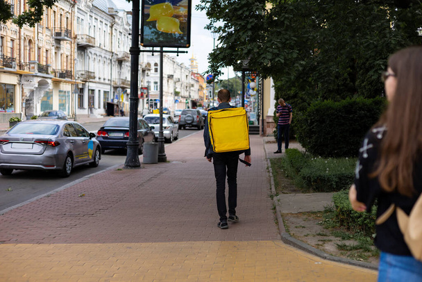 Un joven repartidor caminando con una bolsa térmica amarilla a lo largo de la calle de la ciudad. El repartidor tiene prisa por entregar el pedido. El servicio de entrega viene a entregar rápidamente el pedido  - Foto, imagen
