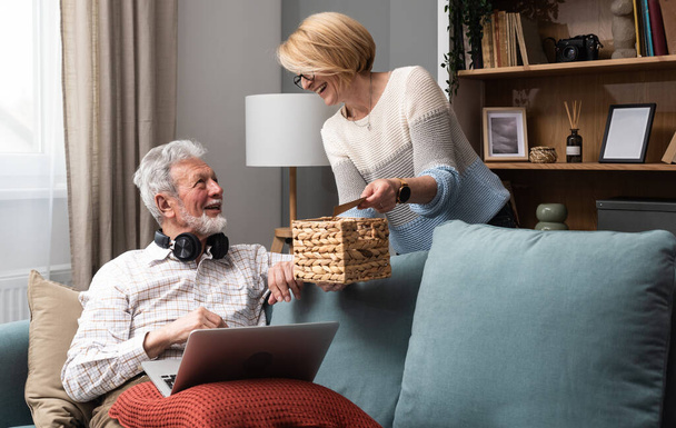 Happy senior couple having fun at their cozy warm home. Elderly man and elderly woman simple living enjoying their retirement with books, laptop, smiling and talking about memories. - Photo, Image