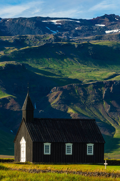 Piękny czarny kościół stoi sam w spokojnym krajobrazie Islandii, otoczony pasmami górskimi i bujną trawą. - Zdjęcie, obraz