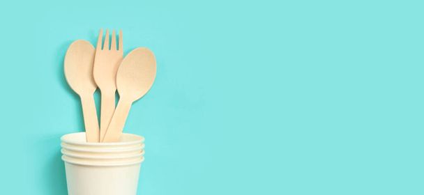 Wooden disposable forks and spoons in a paper cup, top view. Eco friendly disposable kitchen utensils on a blue background, copy space. Flat lay. The concept of ecological dishes - 写真・画像