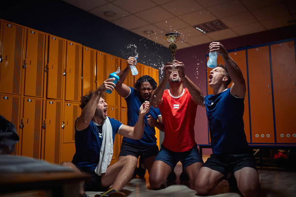 Groep van vier mannen team spelers in de kleedkamer vieren samen succes, houden gouden medaille en schreeuwen van vreugde, spetterend water. Sport, winst, teamconcept. - Foto, afbeelding