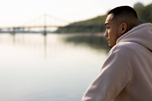 Kısa saçlı bir adam nehrin yanında duruyor ve mesafeye bakıyor. Kapüşonlu genç bir Asyalı bulanık bir nehrin arka planında duruyor. Yüksek kalite fotoğraf - Fotoğraf, Görsel