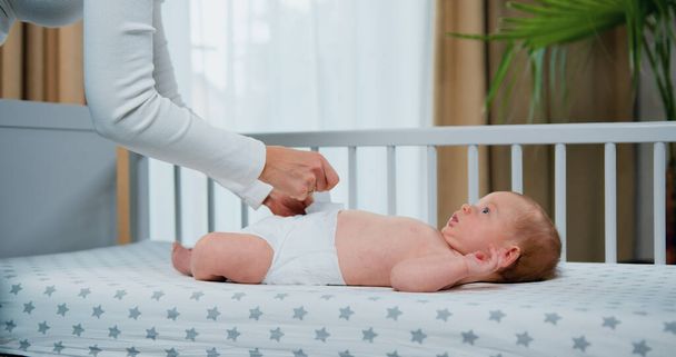 Side view. Mother puts on clean diaper dressing newborn boy lying on bed at home. Close up of moms hands finishing putting clean diaper on infant baby. - Photo, Image