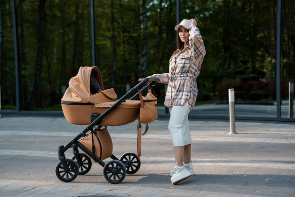 Portrait of a beautiful girl. Close-up photo of a beautiful girl in a plaid shirt and a cap. The girl stands near the brown baby carriage. Mother walking with a small child - Photo, Image