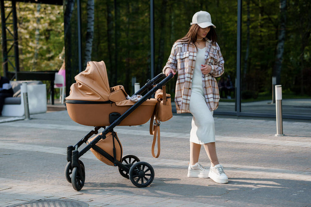 Portrait of a beautiful girl. Close-up photo of a beautiful girl in a plaid shirt and a cap. The girl stands near the brown baby carriage. Mother walking with a small child - Photo, Image