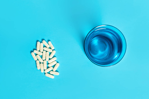 Capsule pills and a blue glass of water on blue background. Top view, fat lay medicine concept. - Foto, Imagen