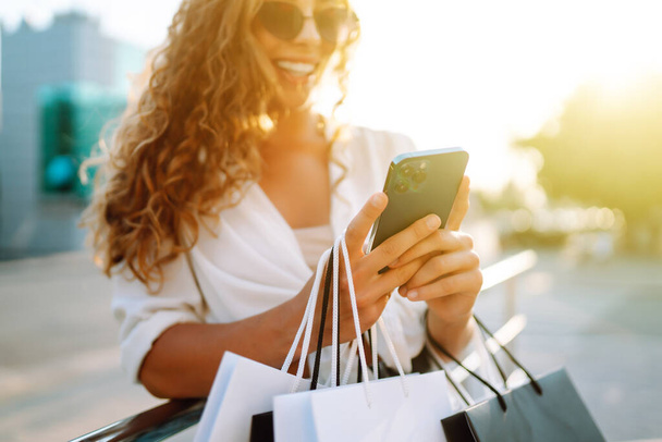 Stylish young woman shopping with a phone on a sunny street. Concept of  online shopping, selling, consumerism. Active lifestyle. - Fotoğraf, Görsel