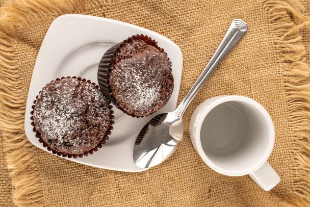 Két házi csokoládé muffin fehér kerámia csészealj csésze, fém kanál és juta szalvéta fa asztalon, makró, felülnézet. - Fotó, kép