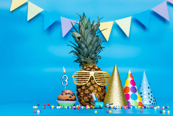 Kreatív háttér ananász karakter napszemüveg másolja a helyet. Boldog szülinapot muffinnal vagy tortával a hármas számú gyertyával. Évforduló ünnep dekorációk egy kék háttér. - Fotó, kép