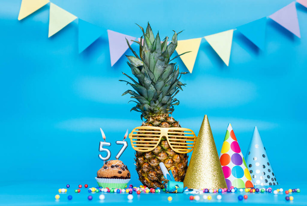 Creative background with pineapple character in sunglasses copy space. Happy birthday background with muffin or cake with candle number  57. Anniversary holiday decorations on a blue background. - Fotó, kép