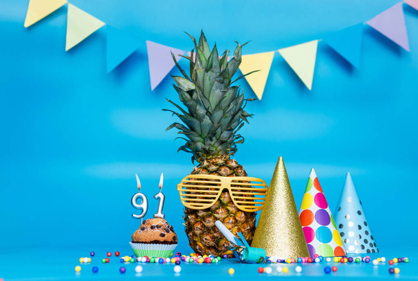 Creative background with pineapple character in sunglasses copy space. Happy birthday background with muffin or cake with candle number  91. Anniversary holiday decorations on a blue background. - Фото, изображение
