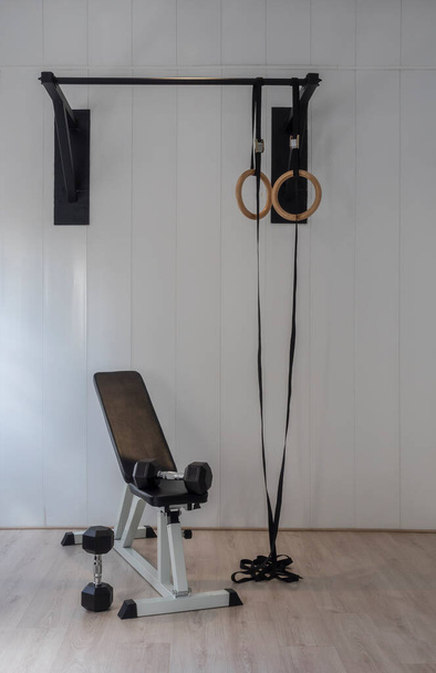 Room without people with rings and pull-up bar in a gym. Dumbbells next to a bench. White background and wooden floor. Healthy lifestyle - Fotografie, Obrázek