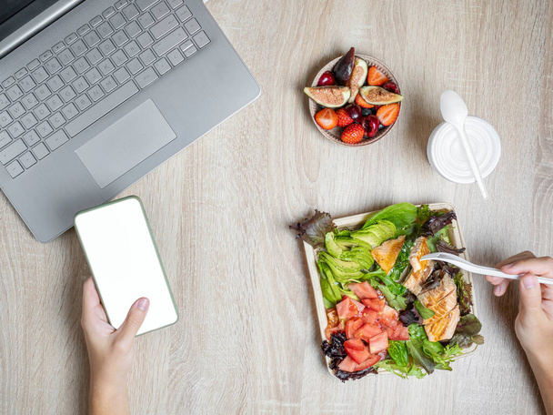 Workspace with laptop, one hand holding a cell phone and the other with a fork eating healthy food takeaway. Lunch takeaway in tapper. View from the top, wooden table in the background. - 写真・画像
