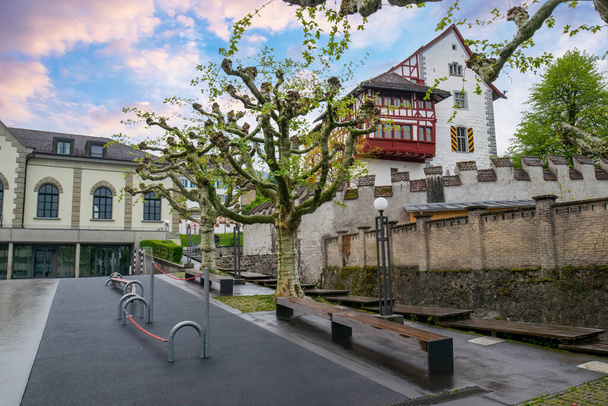 Malerische Straßen im schweizerischen Zug, geschmückt mit einem Fachwerkschloss, dekorativen Bäumen und feuchten Bürgersteigen. Heitere Atmosphäre lädt zum gemütlichen Bummeln ein - Foto, Bild