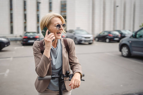 Yetişkin, beyaz, sarışın bir kadın dışarıda elektrikli tekmeyle dikiliyor scooter 'ı itiyor, cep telefonuyla telefon görüşmesi yapıyor şehirde tek başına kendine güveni tam, gerçek insanlar mekanı kopyalıyor. - Fotoğraf, Görsel