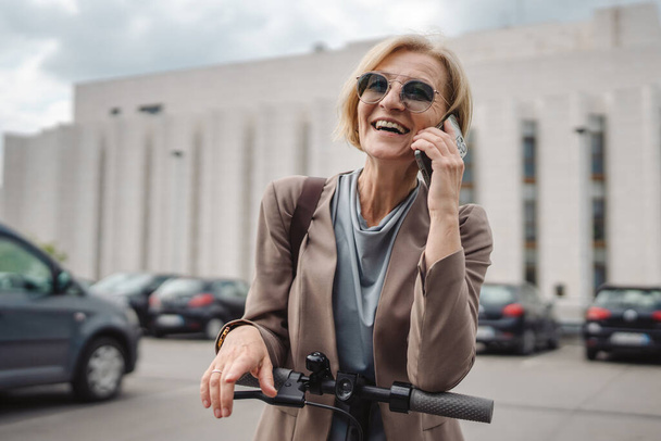 One woman mature caucasian blonde female stand outdoor with electric kick push scooter use mobile phone smartphone to make a call talk in the city stand alone confident real people copy space - 写真・画像