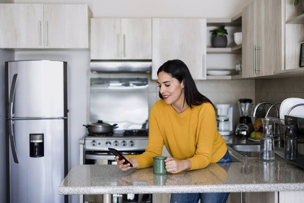 Latin adult woman drinking coffee and using mobile phone in kitchen at home in Mexico Latin America, hispanic female - Фото, зображення
