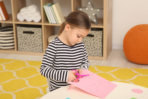 Cute little girl cutting pink paper at desk in room. Home workplace - Photo, Image