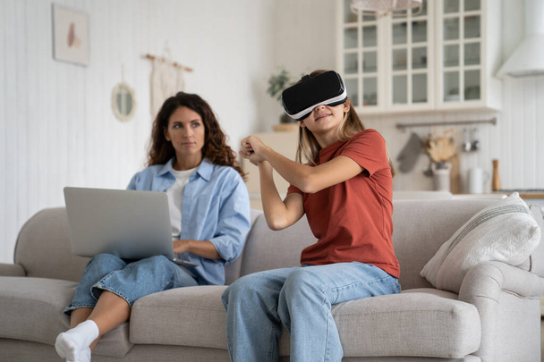 Working from home mother looking at excited little girl daughter wearing VR helmet spending long time immersed in virtual world, selective focus. Dangers of virtual reality for young children - Foto, imagen
