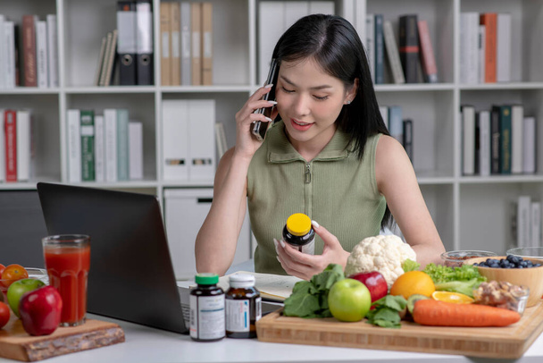 Asian woman professional nutritionist consulting with her client on the phone, surrounded by a variety of fruits, nuts, vegetables, and dietary supplements on the table - Фото, зображення