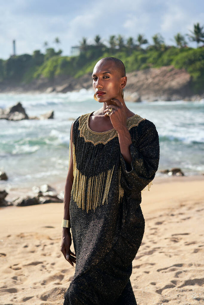 Trans sexual ethnic fashion model in long posh dress and accessories in elegant posture looks at camera. Epatage gay black man in luxury gown poses, touches face on scenic an ocean beach. - Foto, Imagem