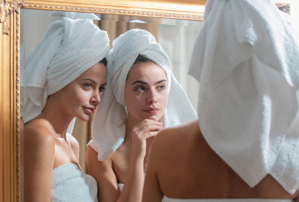 Beautiful woman look in mirror after shower in apartment or hotel room. Young pretty girls friends wrapped in towel smoothing perfecting skin, daily morning routine concept - Zdjęcie, obraz
