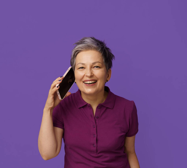 Mature woman wears bright smile in conversation on mobile phone. Isolated on purple background, her grey hair adds air of wisdom and experience, highlighting joy of communication in modern era. High - Фото, зображення