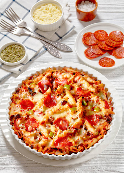 Pizza Casserole of tender penne pasta, browned beef meat, pepperoni, olives, bell peppers, and cheese smothered in marinara sauce in white baking dish on white wood table, vertical view, close-up - Foto, Imagen