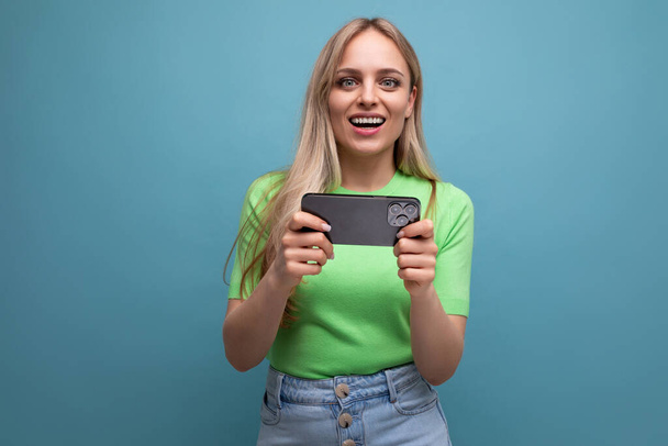 surprised blond girl in casual outfit makes photo using smartphone holding gadget horizontally on blue background. - Φωτογραφία, εικόνα