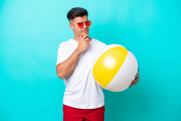 Молодий білий чоловік тримає пляжний м'яч ізольований на синьому фоні, дивлячись в сторону і посміхаючись
 - Фото, зображення