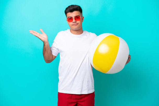 Joven hombre caucásico sosteniendo una pelota de playa aislada sobre fondo azul teniendo dudas mientras levanta las manos - Foto, Imagen
