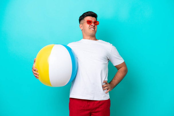 Jonge blanke man met een strandbal geïsoleerd op een blauwe achtergrond die lijdt aan rugpijn omdat hij moeite heeft gedaan - Foto, afbeelding