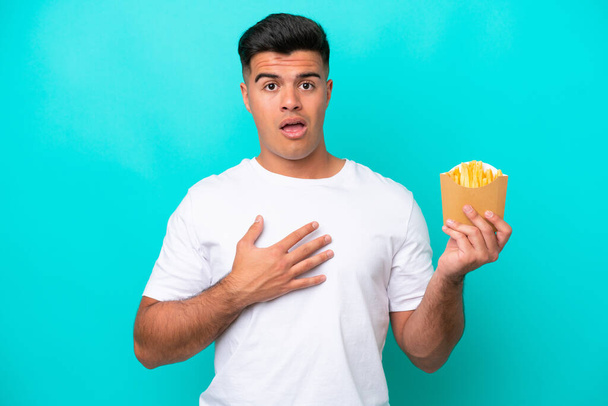 Young caucasian man catching french fries isolated on blue background surprised and shocked while looking right - Photo, Image