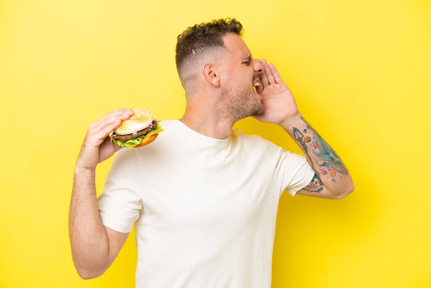 Young caucasian man holding a burger isolated on yellow background shouting with mouth wide open to the side - Photo, Image