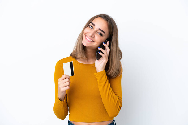 Junge kaukasische Frau isoliert auf weißem Hintergrund hält ein Gespräch mit dem Mobiltelefon und hält eine Kreditkarte - Foto, Bild