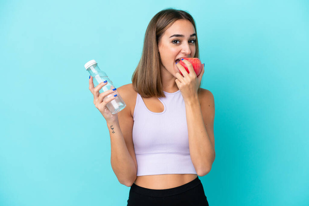 Young caucasian woman isolated on purple background with a bottle of water and eating an apple - Photo, Image