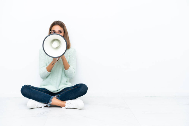 Young caucasian woman sitting on the floor isolated on white background shouting through a megaphone - Photo, Image