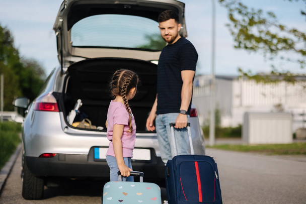 Маленькая девочка несёт чемодан к машине. Папа помогает дочери положить багаж в багажник. Высокое качество фото - Фото, изображение