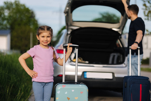 Dad and daughter are standing with their suitcases near a car with an open trunk. Happy little girl preparing to travel . High quality photo - Zdjęcie, obraz